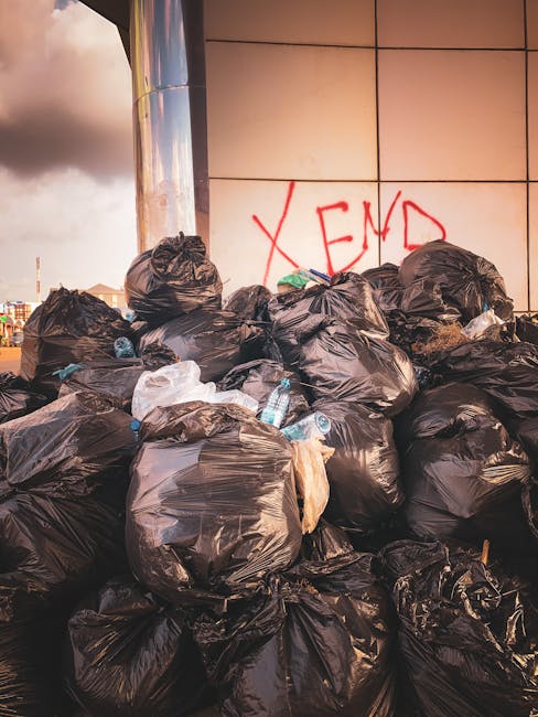 A large pile of black rubbish bags filled with waste sits outside a modern building with a tiled exterior. Several bottles, including plastic water bottles, are visible among the bags, some partially ripped open revealing mixed rubbish inside. The bags are arranged haphazardly, leaning against each other and the building wall. The background shows a cloudy sky and faint outlines of nearby city buildings. On the tiled wall behind the rubbish, there is red graffiti spelling 'XEND.' The scene appears to be an outdoor area designated for waste accumulation, possibly indicating the need for private rubbish collection or on-site clearance services provided by companies like Rubbish Removal Brent. The lighting suggests late afternoon, with a warm glow casting subtle reflections on the building’s surface, emphasizing the texture of the bags and surrounding waste. This setup illustrates a typical scenario where independent waste handling services could be employed as an alternative to standard municipal rubbish collection, supporting safe and efficient rubbish removal in an urban environment.