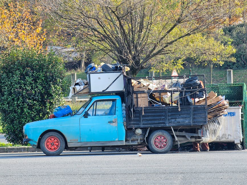 A small, vintage blue pickup truck parked on the side of a road, heavily loaded with a variety of waste materials including large cardboard boxes, black plastic bags, and wooden items stacked in the truck's open cargo area. The truck's wheels are pink, and the body shows signs of wear with visible dirt and minor rust. Behind the truck, there is a green metal fence and a large deciduous tree with yellow-green leaves, suggesting an autumn setting. Nearby, a small leafy shrub is visible on the roadside. The scene appears to be during daylight with natural lighting illuminating the surroundings, illustrating an example of private or independent rubbish removal activity in a residential or semi-urban area, supported by local waste management services like Rubbish Removal Brent.