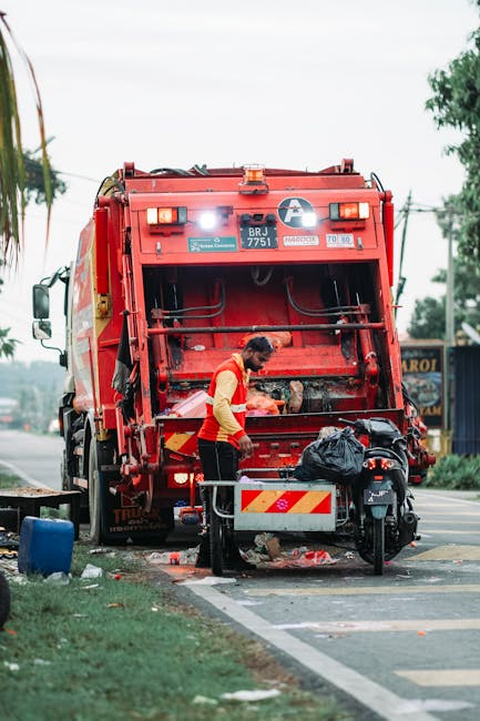 A waste collection worker in a yellow and red uniform is standing at the rear of a large, bright red garbage truck, which is parked on the side of a street near a curb. The back of the truck is open, revealing hydraulic mechanisms and storage areas for rubbish. The worker appears to be handling debris or waste materials, with a trash bag visible nearby on top of a small mobile waste container attached to the truck. The scene is set outdoors with a background of trees, utility poles, and a building, under an overcast sky. The surrounding paved road, curb, and a patch of grass indicate an urban or suburban environment typical for rubbish removal services, supporting the context of private waste management or alternative collection. The truck's reflective stripes and safety equipment are visible, highlighting operational safety. This image reflects a waste collection process potentially managed independently by Rubbish Removal Brent, fitting into the context of Neasden estate rubbish collection or private disposal activities.