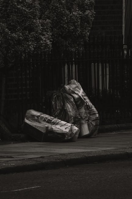 A pair of large black plastic garbage bags filled with waste are positioned on the pavement beside a black metal fence with vertical bars. The bags appear to contain household rubbish and are partially crumpled, showing a shiny, reflective surface. Behind the fence, there is foliage from dense bushes or trees, and a dark structure, possibly a building, is visible in the background. The scene is illuminated with natural light, creating shadows on the street and the bags. The setting suggests an area awaiting collection for municipal or private rubbish removal, aligning with on-site waste clearance services provided by companies like Rubbish Removal Brent. The environment appears calm and clean, with no other objects or debris visible near the bags, indicating an organized approach to waste disposal within an urban or residential area, consistent with private rubbish removal methods.