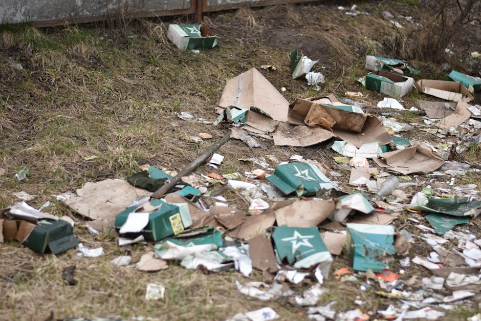 A patch of ground with scattered litter, including torn cardboard boxes, crumpled paper, and discarded packaging materials, predominantly in shades of brown, green, and white. Some cardboard boxes, originally green with white logos, are flattened or partially crushed, and debris such as plastic bottles and paper fragments are mixed among the boxes. The litter is spread across a grassy area with patches of bare soil and overgrown vegetation in the background. In the top left, a wooden fence is visible, with parts of the grass appearing uneven and slightly muddy, indicating a neglected outdoor space. The overall scene appears to be an informal dumping site, with waste items randomly strewn on the ground, highlighting the need for professional rubbish removal services such as those offered by Rubbish Removal Brent, who specialize in on-site clearance and private waste handling solutions for properties in the Wembley Park NW10 area.