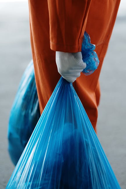 Close-up of a person wearing orange overalls and a white glove, holding a tightly tied blue plastic rubbish bag with a shiny surface that reflects light. The individual is standing outdoors on a paved surface, with a slightly blurred background suggesting an open area or street scene. The bag appears to contain waste materials, possibly household or collection debris, and is positioned in the foreground, emphasizing the act of rubbish collection or disposal. The scene reflects a typical scene of waste handling, relevant to independent rubbish removal services, such as those offered by Rubbish Removal Brent, indicating the process of private waste collection and transfer to disposal facilities. The lighting is even, with natural daylight illuminating the scene, highlighting textures of the plastic bag and the fabric of the overalls, conveying a professional, observant perspective suitable for a waste management context.