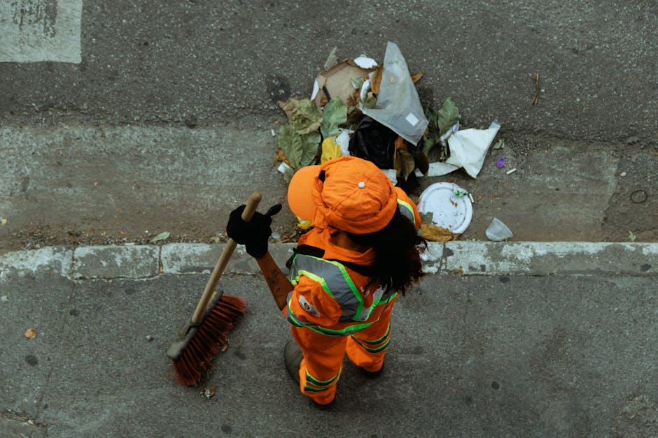 A street-level view showing a street cleaner wearing an orange high-visibility uniform with reflective stripes, an orange cap, black gloves, and sturdy boots, standing on a pavement beside a concrete wall. The worker is holding a long-handled brush with a wooden handle and red bristles, and is facing a pile of mixed waste that includes crushed paper, cardboard, plastic bags, and food packaging, scattered along the edge of the wall. The area appears to be part of a dedicated waste collection or street cleaning activity, with the debris accumulated at the curbside. The environment is lit with natural light, and the scene captures an outdoor urban setting focused on waste management tasks that support rubbish removal services such as those offered by Rubbish Removal Brent.